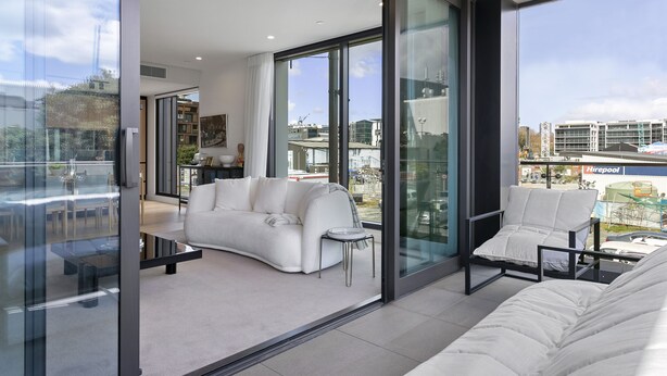 Un salón moderno con un sofá blanco, una mesita y una silla, grandes ventanales con vistas a la ciudad.