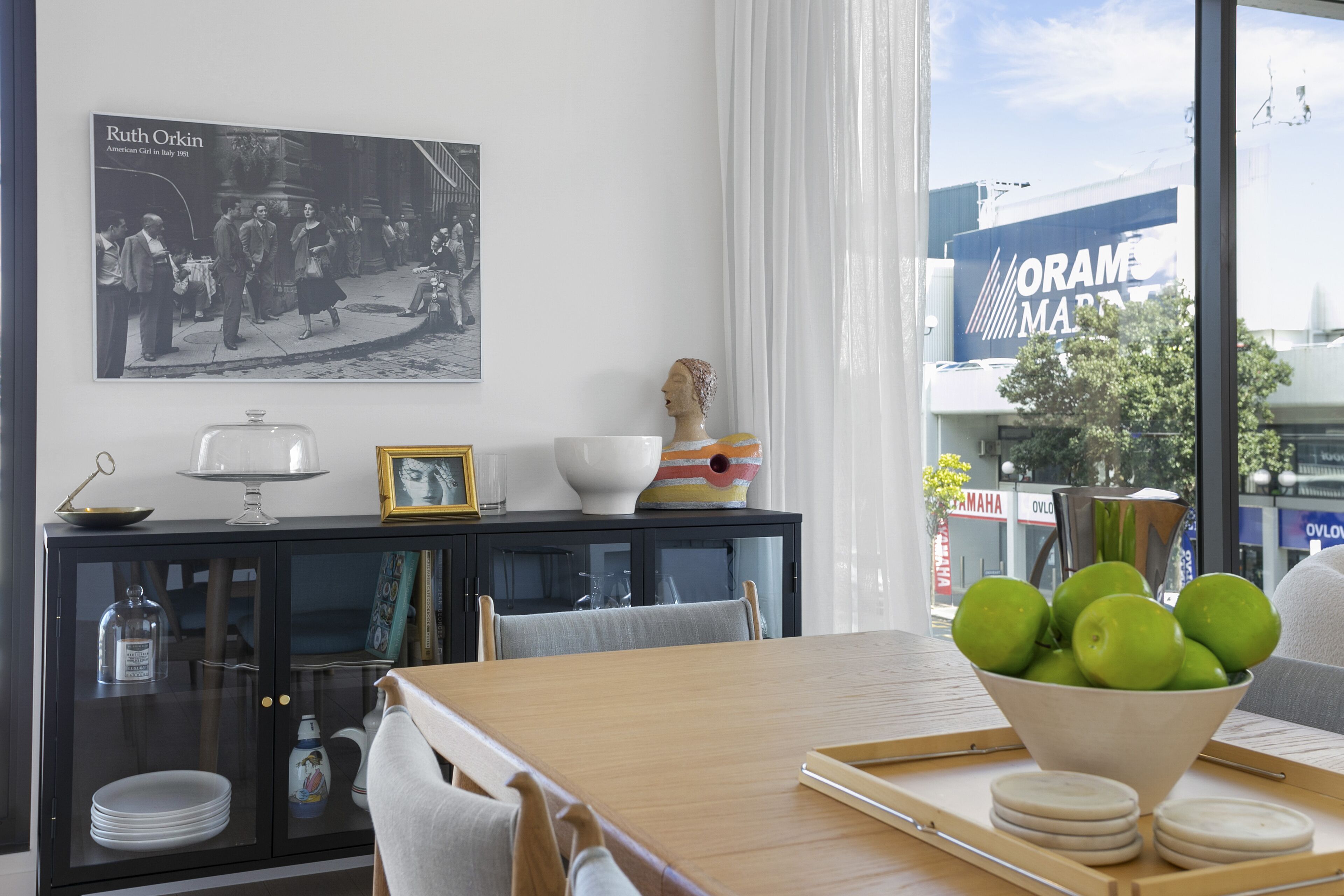 A dining area with a wooden table, a bowl of green apples, a black cabinet with glass doors, and a framed photograph on the wall.
