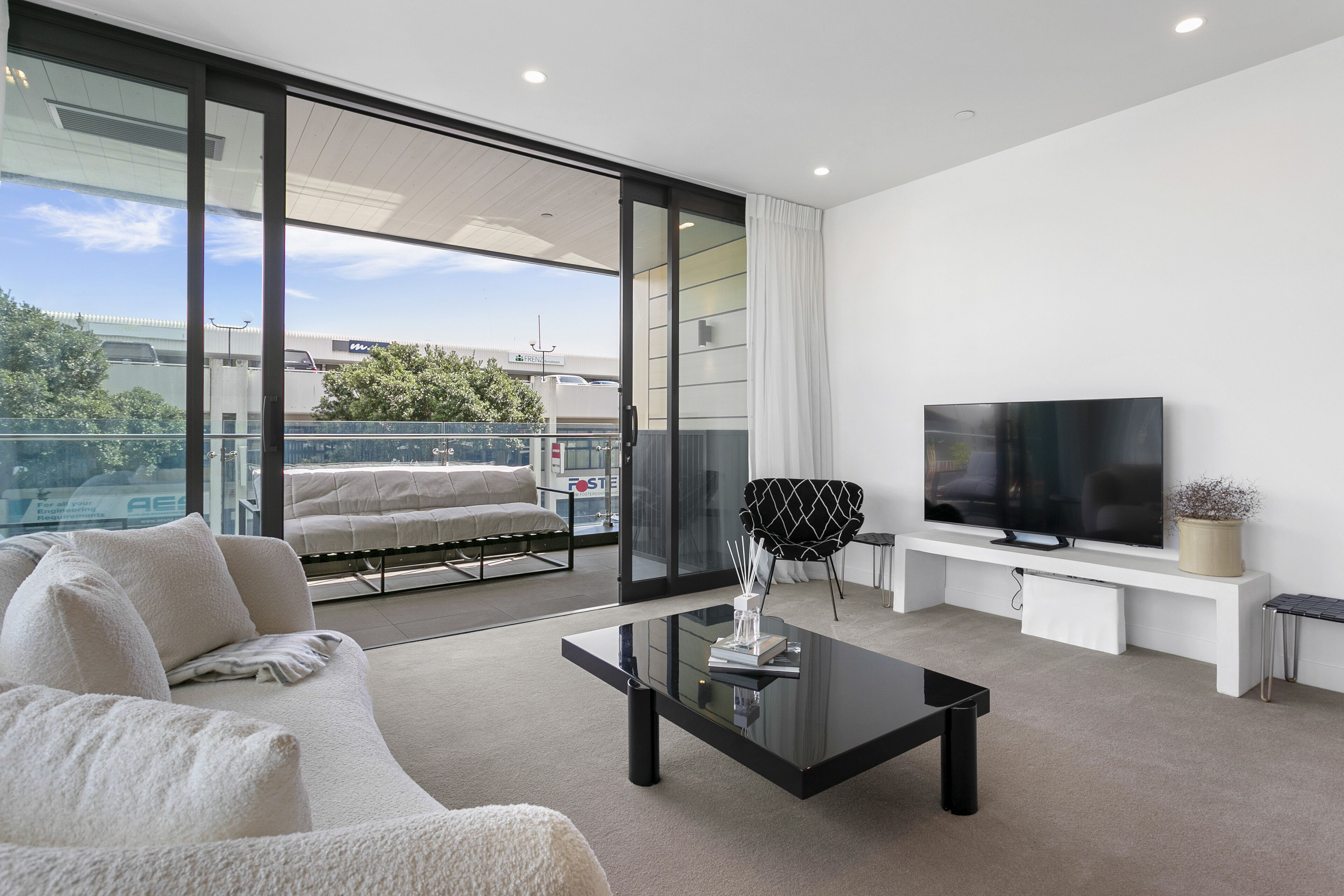 A modern living room with a sofa, a glass coffee table, a TV on a white stand, and a balcony with a view of the city.