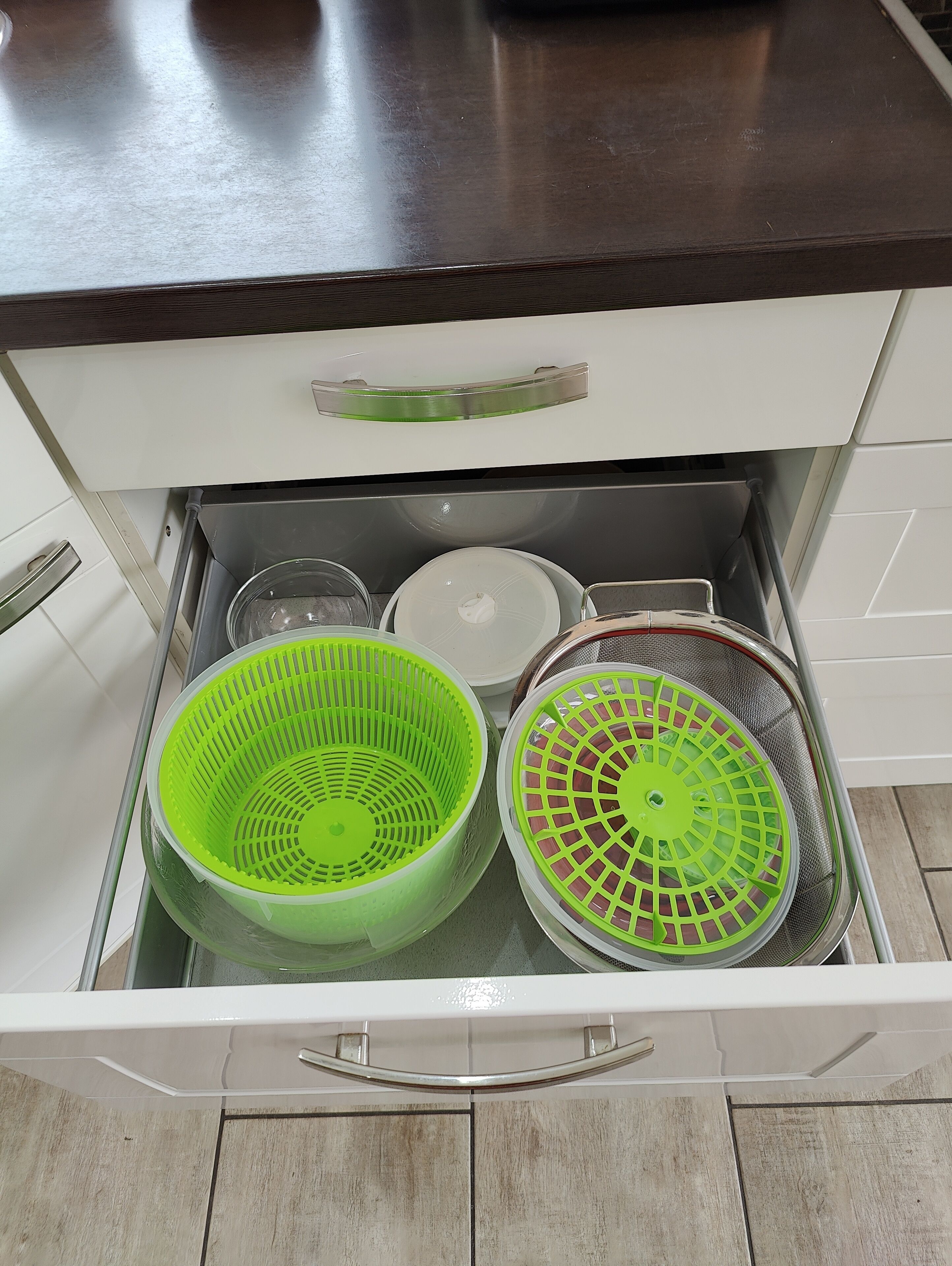 A kitchen drawer with a white handle, containing a green colander, a white colander, and a metal strainer basket.