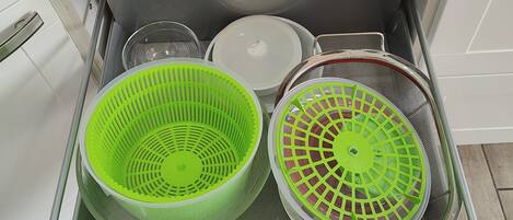 A kitchen drawer with a white handle, containing a green colander, a white colander, and a metal strainer basket.