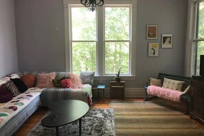 A cozy living room with a sofa, a small round table, a bench, and a large window with a view of trees. - Lovely Victorian near Bathhouse Row (Hot Springs)