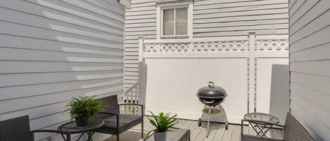 Une terrasse couverte, aménagée avec du mobilier d’extérieur, un barbecue et une clôture blanche.