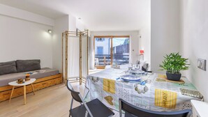 En moderne stue med en sofa, et spisebord dækket til to personer og et stort vindue med udsigt til bygninger.
