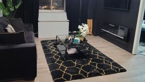 Un saló modern amb un sofà negre, una taula de centre de vidre, una catifa negra i daurada, un televisor de pantalla plana penjat a la paret i una finestra amb cortines.