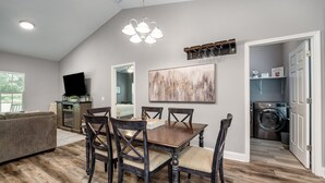 A dining area with a wooden table and chairs, a painting on the wall, and a view into the kitchen. - Cozy House in the Woods - 25 minutes from downtown (Pegram)