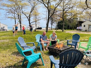 A group of people sitting around a fire pit in a grassy area with chairs and a table, surrounded by trees and a clear sky.