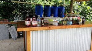 Zona de bar con varias botellas y recipientes, un lavamanos y barriles azules al fondo.