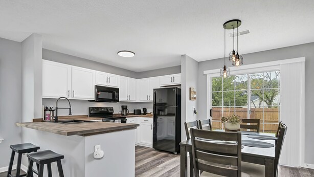 A modern kitchen with white cabinets, a central island with stools, a dining area with a table and chairs, and a view of the backyard through large windows. - NEW! Beautiful Updated House w/ Fenced Backyard! (Columbia)