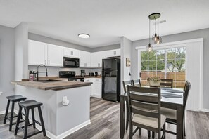 Una cocina moderna con armarios blancos, una isla central con taburetes, un comedor con mesa y sillas, y vistas al patio trasero a través de amplios ventanales.
