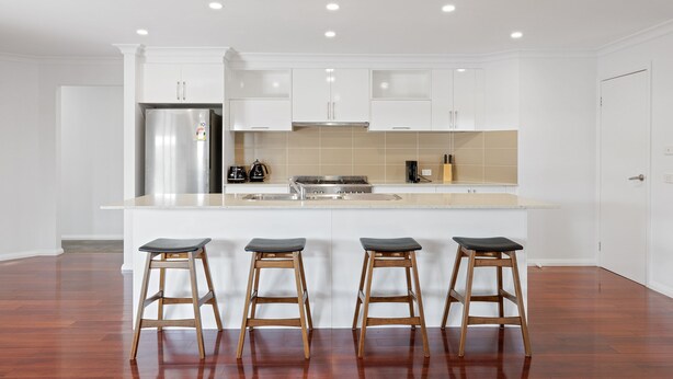 Una cocina moderna con armarios blancos, un refrigerador de acero inoxidable, una estufa a gas y tres taburetes de madera.