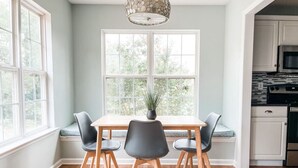 Un coin repas avec une table en bois et quatre chaises, une fenêtre donnant sur un paysage verdoyant, et une cuisine visible à travers une porte ouverte.