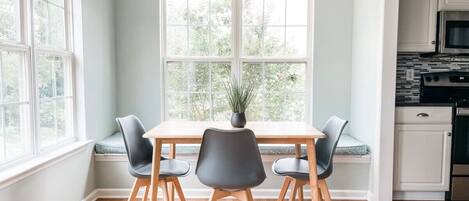 A dining area with a wooden table and four chairs, a window with a view of greenery, and a kitchen visible through an open doorway.
