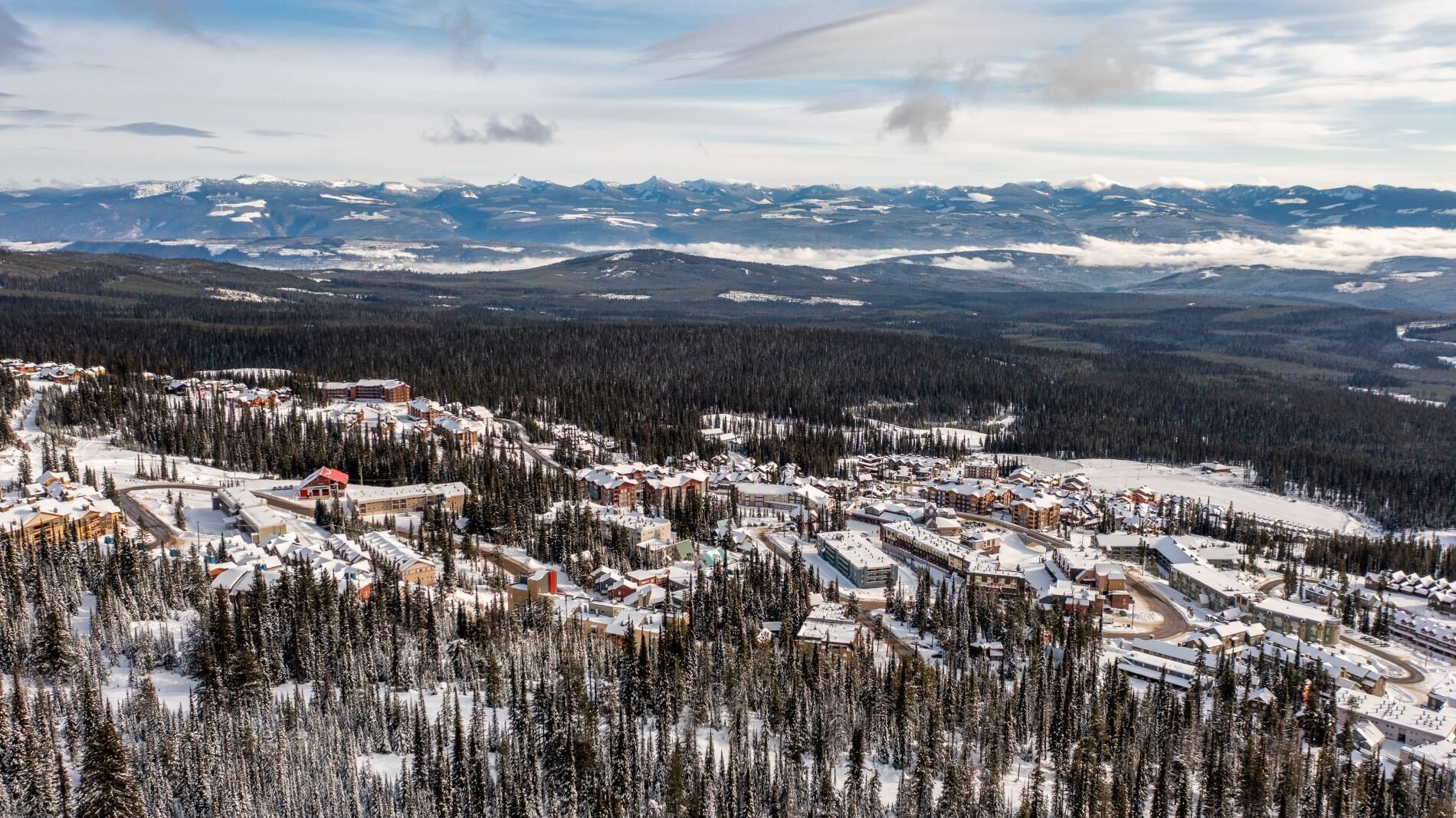 Ein schneebedecktes Resort mit mehreren Gebäuden, umgeben von dichten Wäldern und Bergen im Hintergrund.