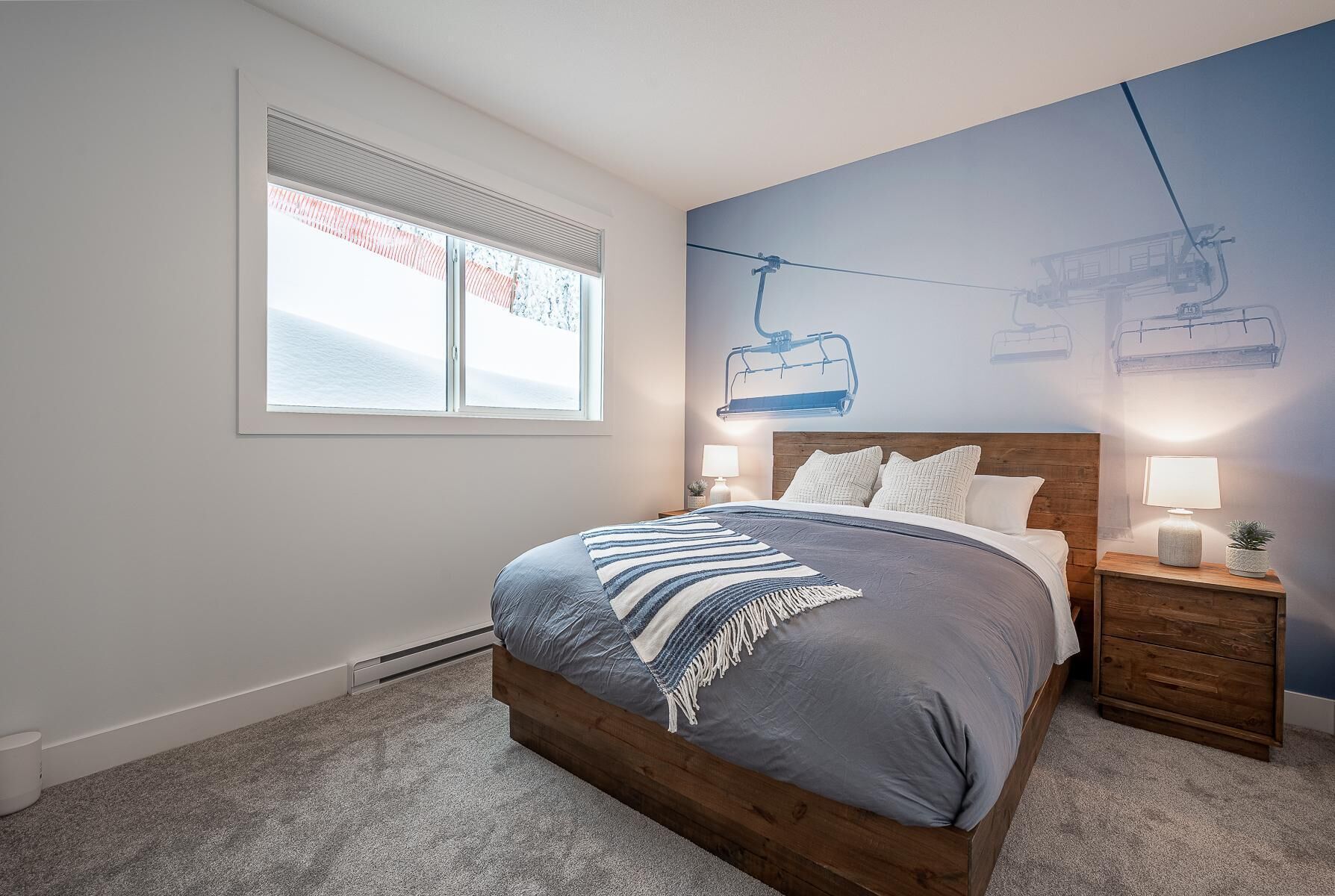 A bedroom with a large bed, a window with blinds, a wall mural of a ski lift, and a nightstand with a lamp.