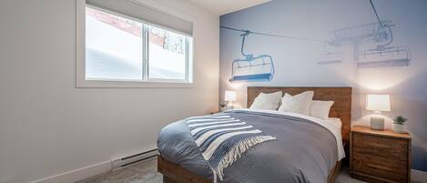 A bedroom with a large bed, a window with blinds, a wall mural of a ski lift, and a nightstand with a lamp.