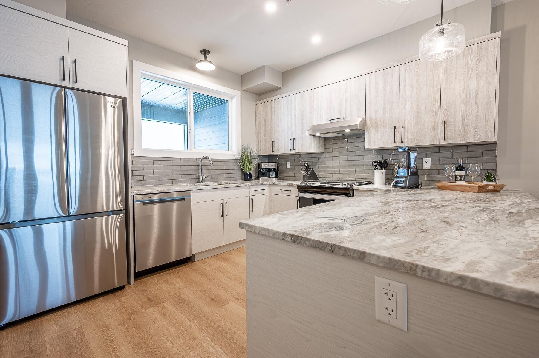 A modern kitchen with stainless steel appliances, light wood cabinetry, a marble countertop, and a large window providing natural light.