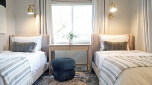 A hotel room with two single beds, a small desk, a potted plant, and a window with curtains.