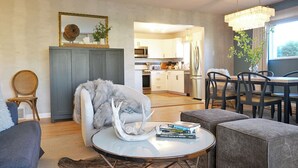 A modern living room with a grey sofa, a round coffee table with books, a fur throw, and a decorative antler. A dining area with a chandelier and a view into the kitchen is visible in the background.