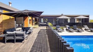 Et moderne poolområde med liggestole og parasoller, træterrasse og udsigt til en moderne bygning.