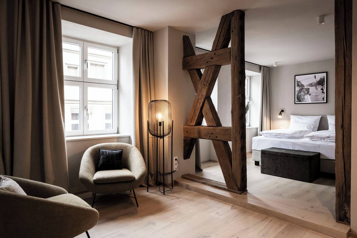 Ein Hotelzimmer mit großem Fenster, einem Bett mit weißer Bettwäsche, einer Holzbalkenkonstruktion, einem Sessel mit Kissen und einer Stehlampe.