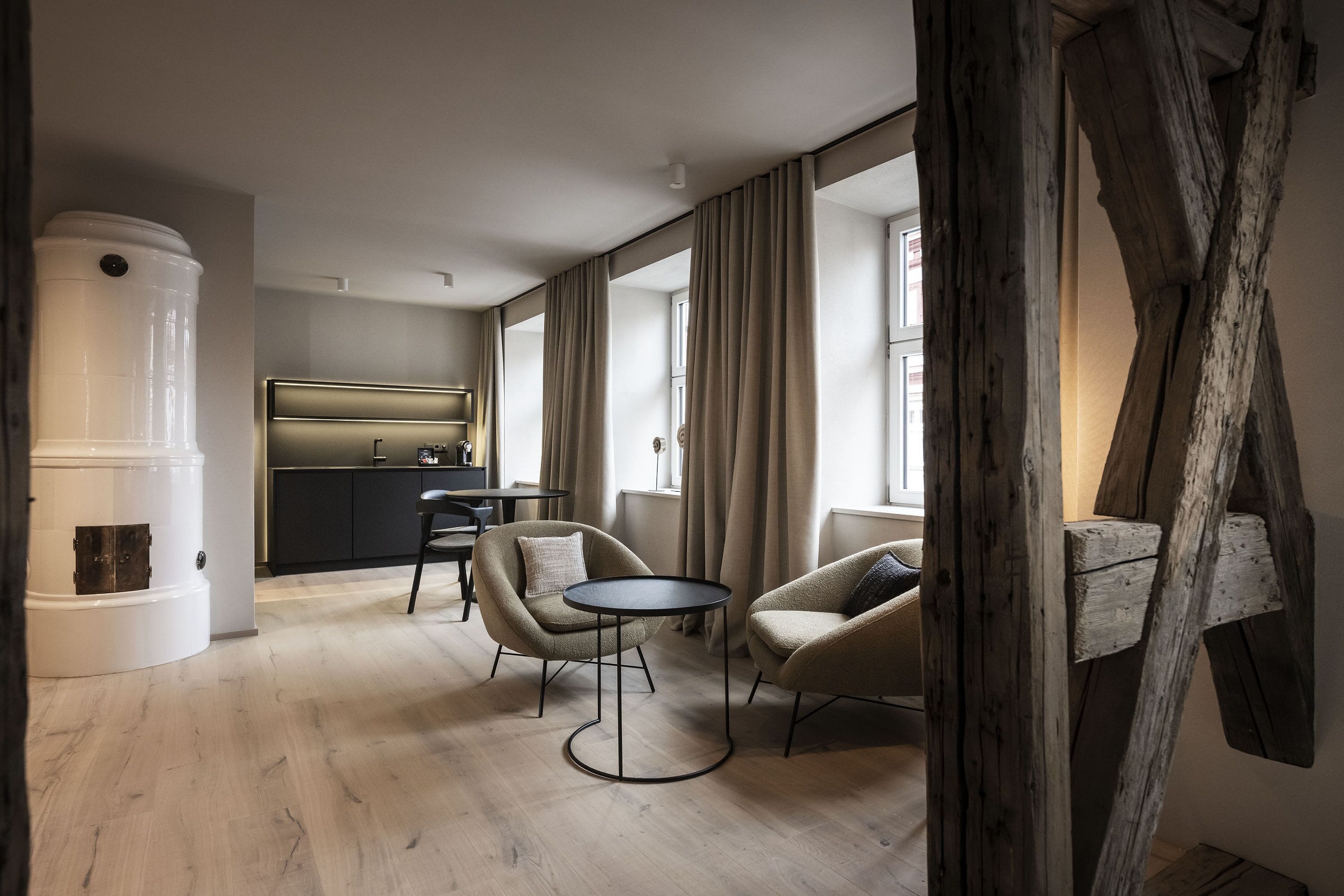 Ein modernes Wohnzimmer mit zwei Sesseln und einem kleinen runden Tisch, im Hintergrund eine Küche und ein großes Fenster mit Vorhängen.