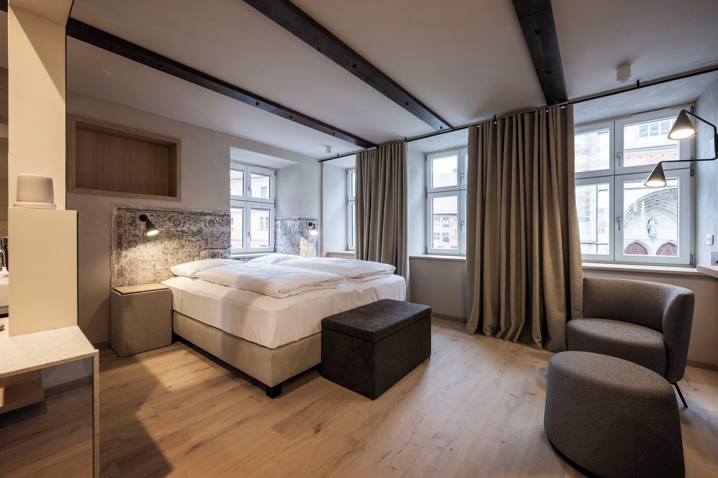 A modern bedroom with a large bed, bedside tables, a chair, and a view of the city through large windows.