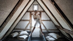 Una persona de pie en un loft con techo inclinado, vigas de madera y un ventanal pequeño. Se ven almohadas y la estructura de una cama.