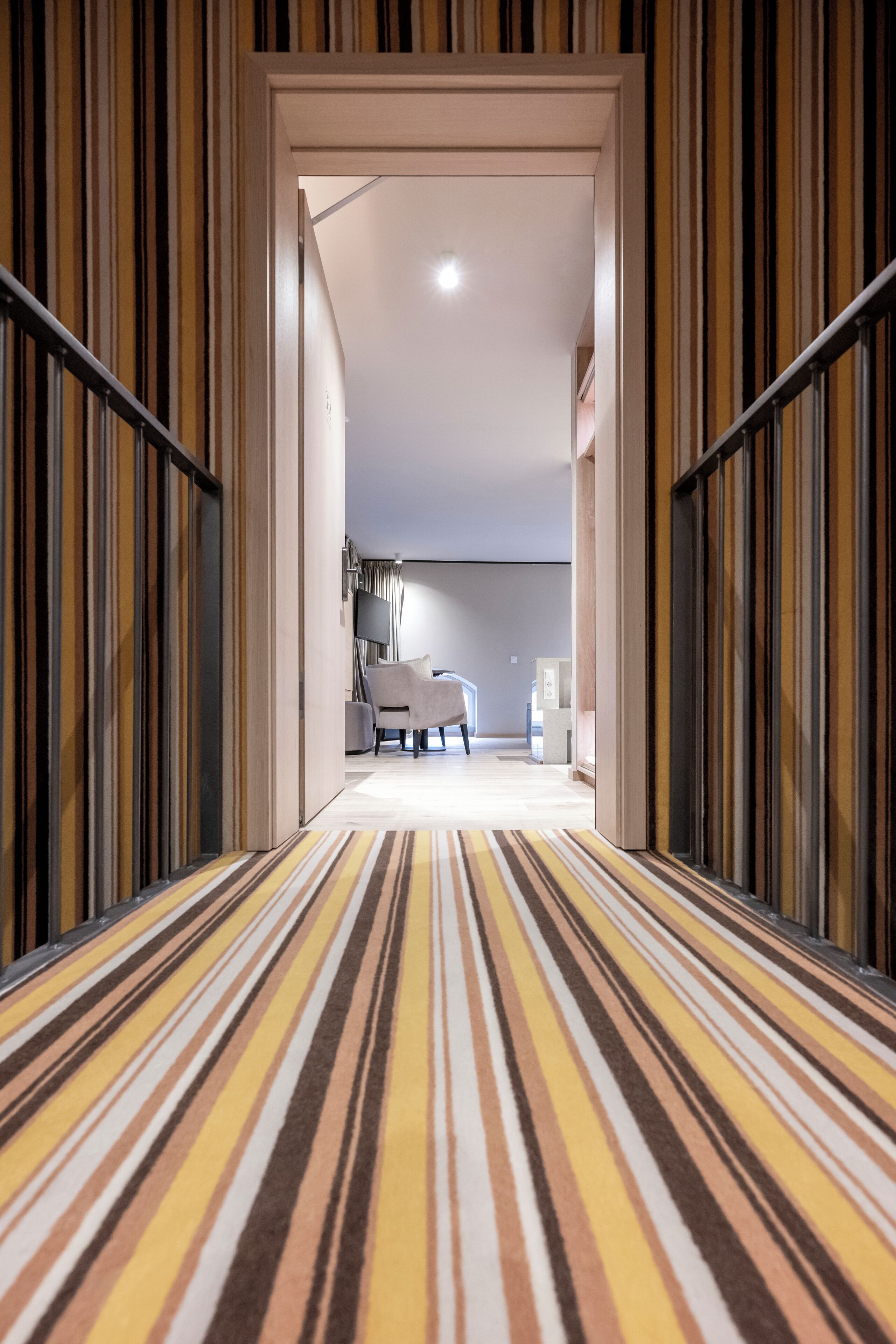 A hallway with striped carpet leading to a room with a chair and a flat-screen TV.