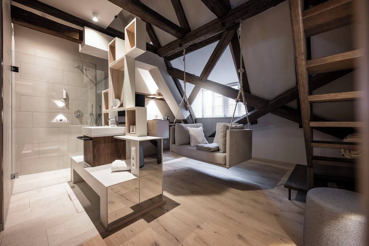 Ein modernes Schlafzimmer im Loft-Stil mit schrägverdeckter Decke, eingebauten Holzregalen, einer Duschkabine aus Glas und einer gemütlichen Sitzecke mit Sofa und Kissen.