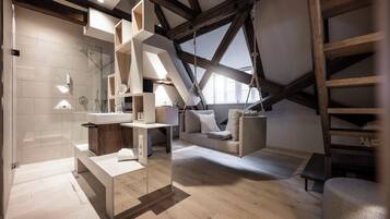 A modern loft bedroom with a sloped ceiling, built-in wooden shelves, a glass shower enclosure, and a cozy seating area with a sofa and cushions.