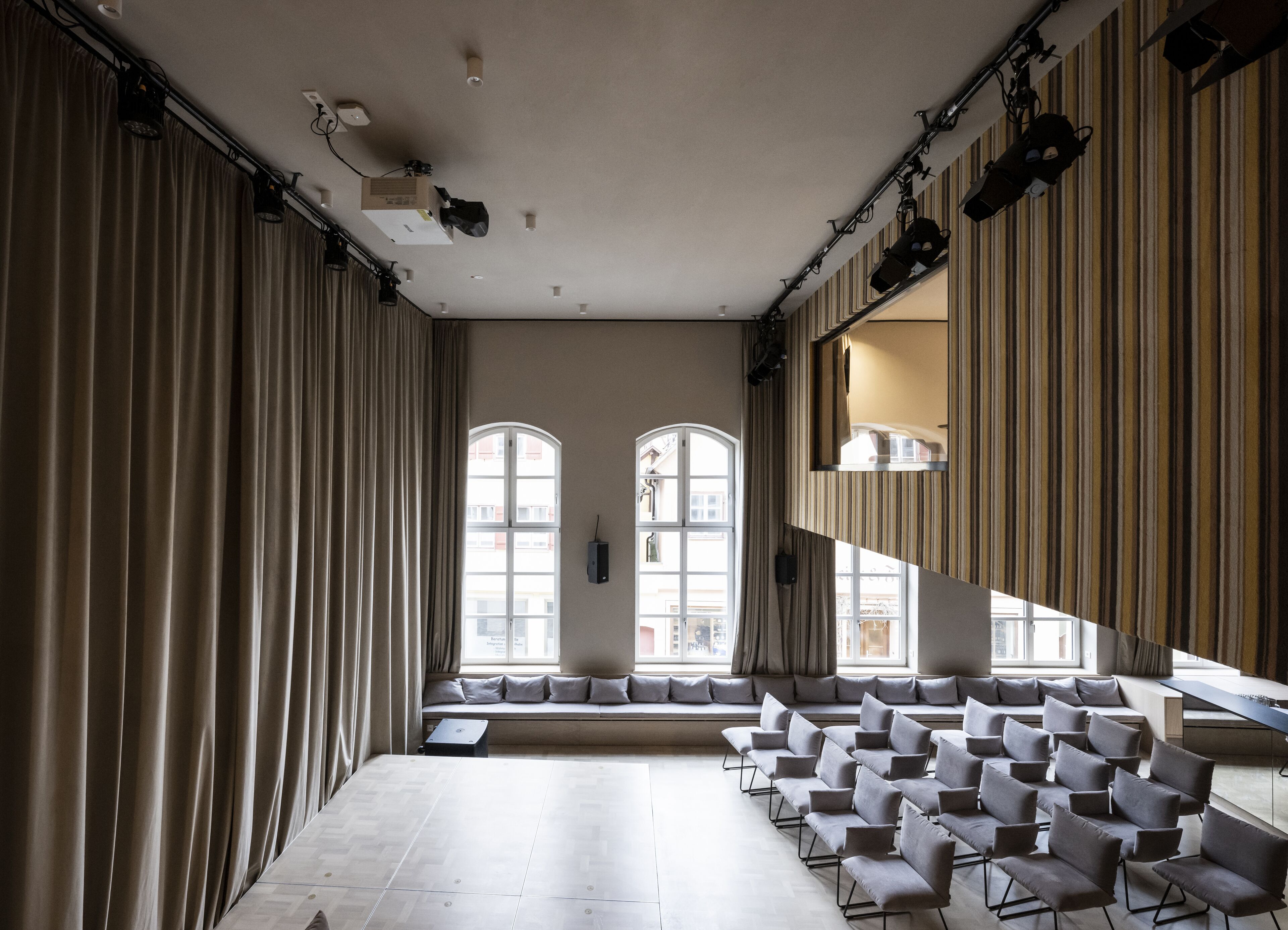 A conference room with a long table and rows of chairs, large windows with curtains, and stage lighting.
