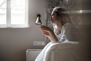 Eine Person sitzt auf einem Bett in einem Zimmer mit Fenster und einer Wandleuchte.