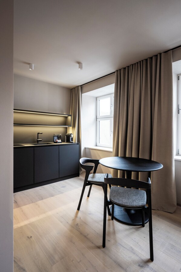 A modern kitchen with a dark countertop, a sink, and a coffee machine. There is a round table with two chairs and a window with curtains.