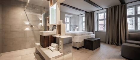 A modern bathroom with a shower, vanity, and a large mirror. There is a bed and a window with curtains in the adjacent room.