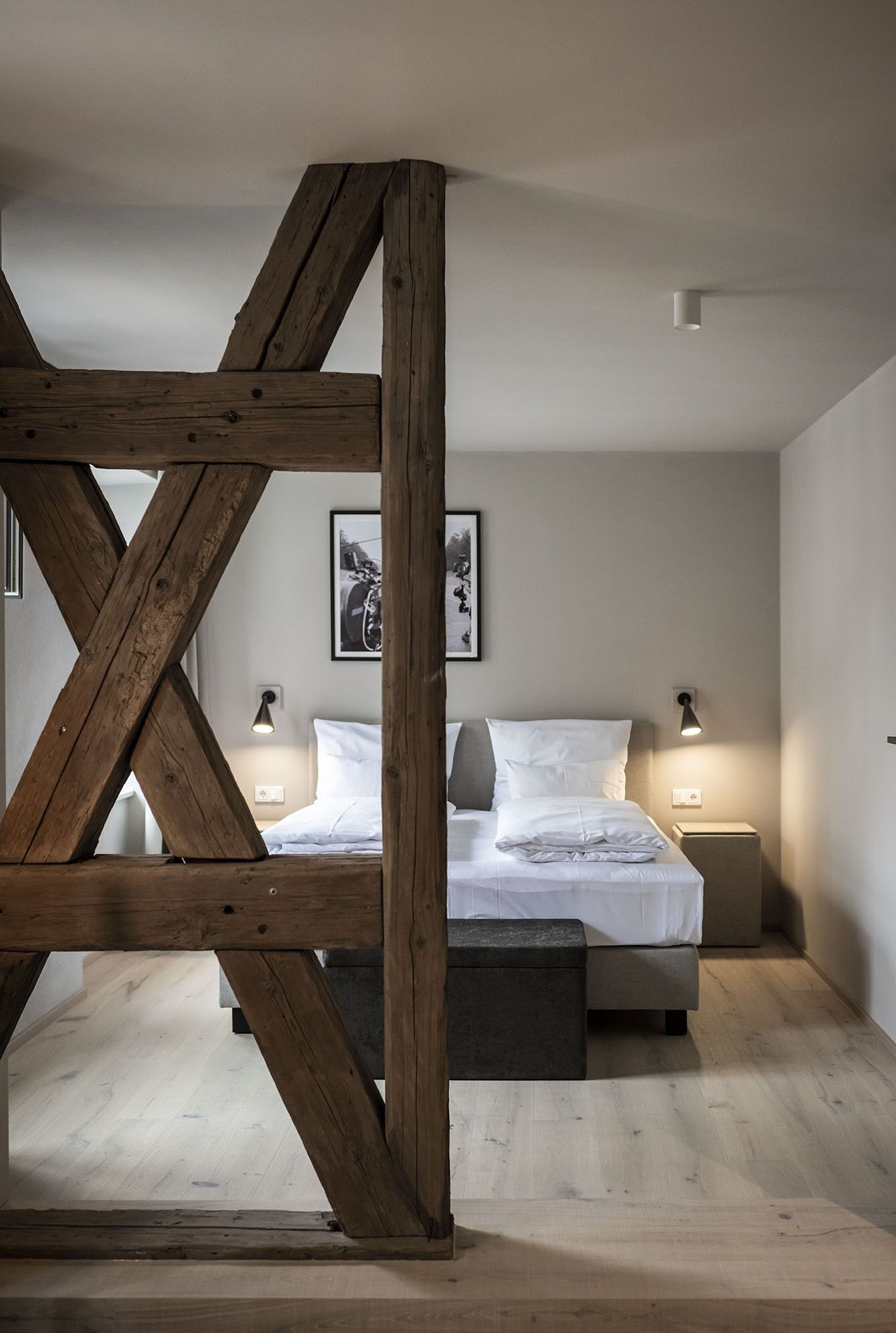 A bedroom with a wooden truss ceiling, a bed with white bedding, two bedside tables with lamps, and two framed pictures on the wall.
