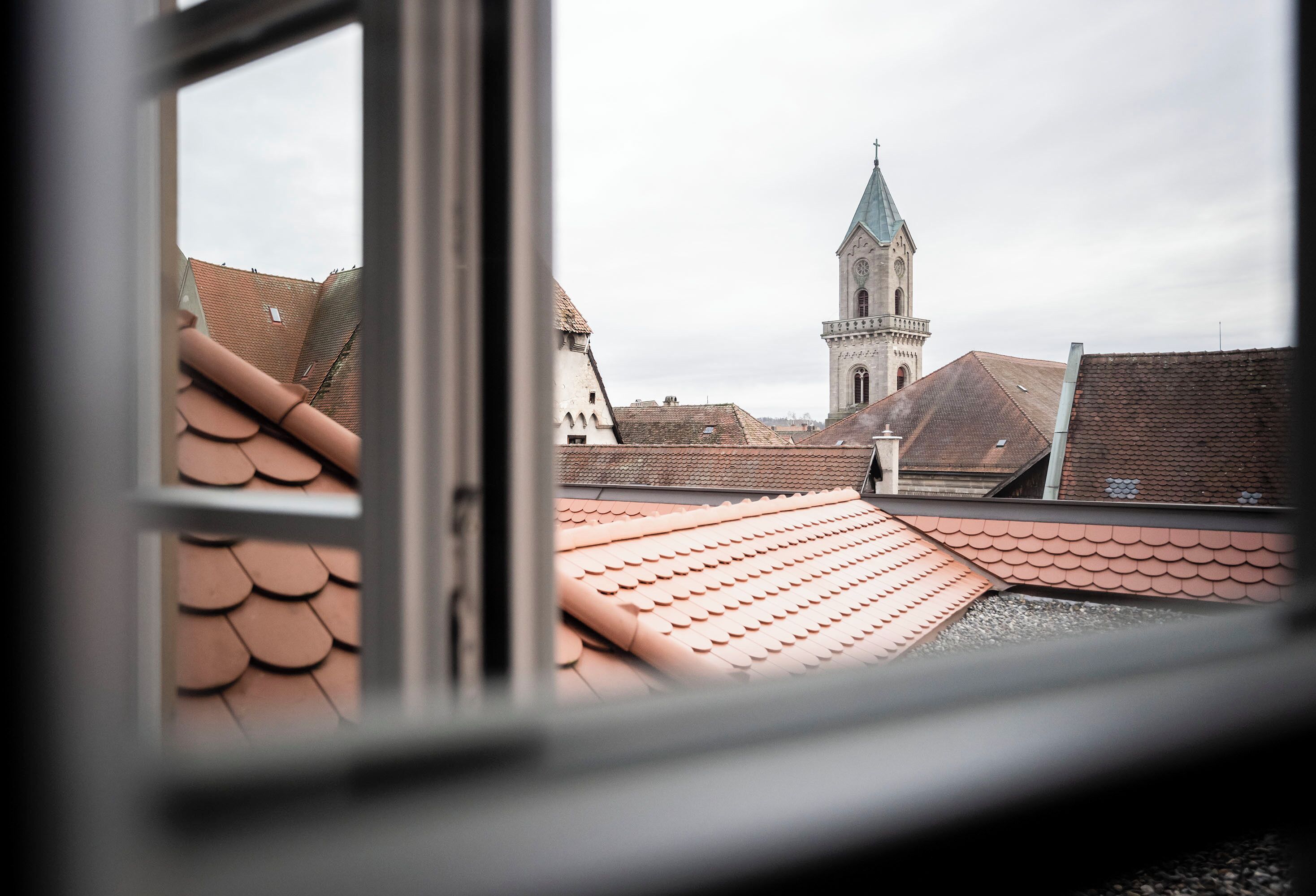 A view through a window of a building with terracotta roofs and a church with a spire in the background.