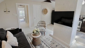 A living room with a black sofa, a patterned rug, a woven stool, a white chair, a flat-screen TV, and a fireplace.