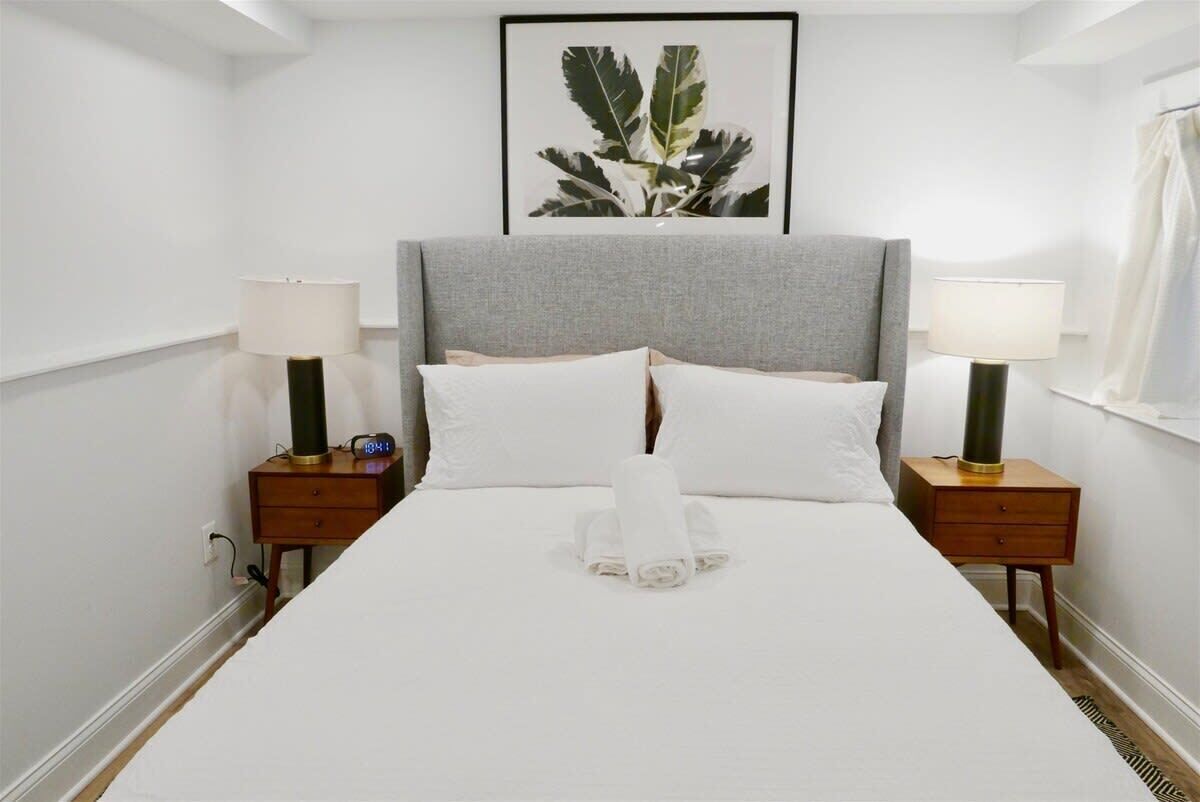 Een netjes opgemaakt bed met witte lakens, twee nachtkastjes met lampen, en een lijst met een plant aan de muur.
