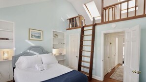 Une chambre à coucher avec un lit, une échelle, une lucarne, une bibliothèque et une porte donnant sur une autre pièce.
