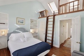 Un dormitorio con cama, escalera, tragaluz, estantería y puerta que da a otra habitación.