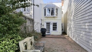 Un étroit sentier pavé de briques mène à une maison blanche dotée d’une baie vitrée et d’un espace barbecue abrité.