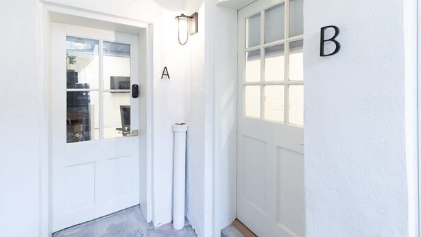 A white exterior of a building with two doors labeled A and B, a light fixture above door A, and a small white pipe near the door.