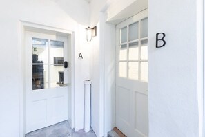 Een wit gevel van een gebouw met twee deuren, genummerd A en B, een lamp boven deur A, en een kleine witte pijp in de buurt van de deur.
