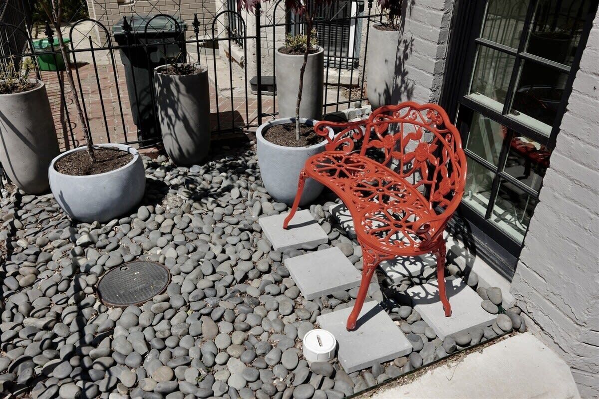 Un espace patio avec un banc en métal rouge, des plantes en pot et une grille métallique au sol.