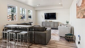A modern living room with a sectional sofa, a dining table with bar stools, a flat-screen TV mounted on the wall, and a large window offering a view of the outdoors. - Rustico Roost by RedAwning (Washington)