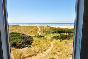 Vista de una playa con un sendero arenoso que conduce a la orilla, rodeada de dunas cubiertas de hierba y vegetación.