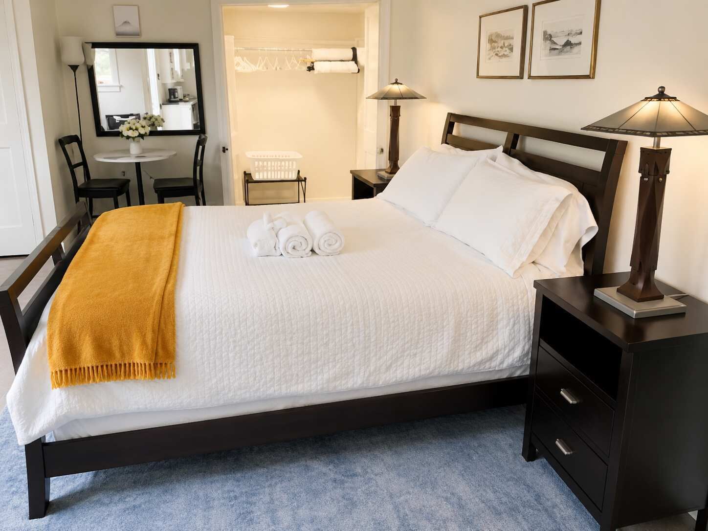 Un lit bien fait, avec une couette blanche, un plaid de couleur moutarde et des serviettes pliées. Une table de chevet en bois foncé, sur laquelle se trouve une lampe, est placée à droite.