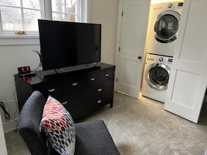 Ein kompakter Wohnraum mit einem Flachbildfernseher auf einer schwarzen Kommode, einem grauen Sofa mit einem gemusterten Kissen und einer Frontlader-Waschmaschine.