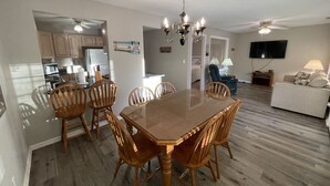 A spacious living room with a wooden dining table and chairs, a ceiling fan, a flat-screen TV, and a sofa.
